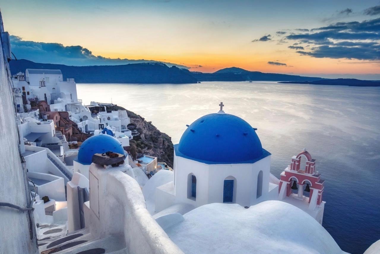 A blue-domed church and pink campanile above the caldera at dusk in Oia